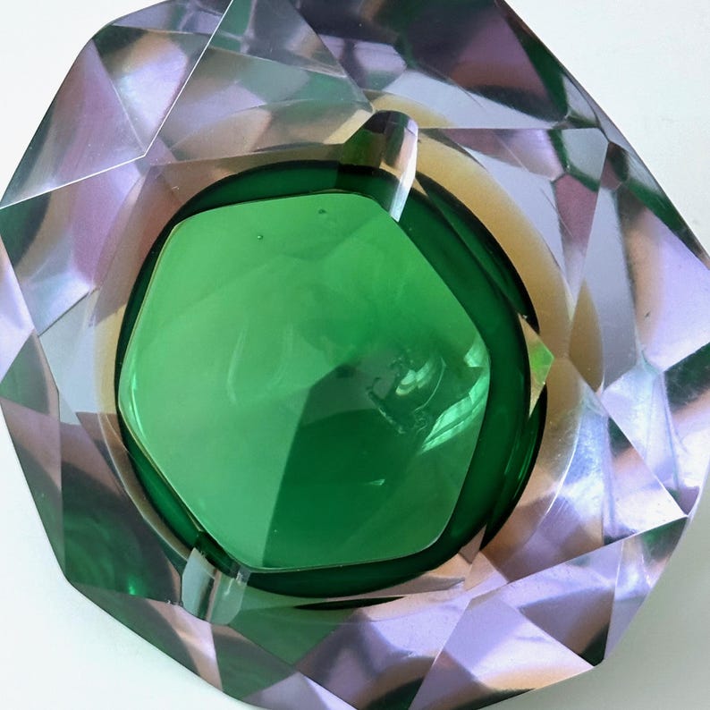 Op de afbeelding: Een facetgeslepen, smaragdgroene asbak met een centrale kom. De asbak heeft een geometrisch ontwerp met een heldere, reflecterende buitenkant. De binnenkant is diepgroen, wat een opvallend contrast cre&euml;ert. De algemene vorm is een veelzijdig veelhoek.