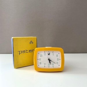 Könnte beinhalten: Ein gelber Vintage-Wecker mit weißem Zifferblatt und schwarzen Zeigern, neben der Originalverpackung. Das Zifferblatt zeigt die Uhrzeit, und auf der Box steht der Name "peter".