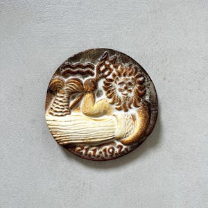 May include: A round, brown and cream-colored ceramic relief with a detailed design. The relief depicts a reclining figure, a sun, and other stylized elements. The date "27.1.192" is inscribed at the bottom. The overall design has a vintage aesthetic.
