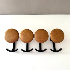 May include: A set of four wall hooks, each featuring a circular light brown wooden knob with a visible grain pattern attached to a curved black metal base. The hooks are arranged in a horizontal row, showcasing the contrast between the natural wood texture and the smooth, dark metal finish of the functional hardware components.