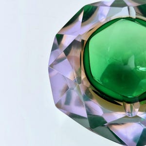 Op de afbeelding: Een facetgeslepen asbak van smaragdgroen en helder glas. De asbak heeft een geometrisch ontwerp met een centrale groene kom en een heldere, diamantgeslepen buitenkant. De randen zijn schuin, waardoor een spel van licht en schaduw ontstaat.