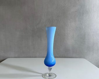May include: A tall, elegant blue glass vase with a flared top and a bulbous base. The vase features a gradient of blue hues, transitioning from a lighter shade at the top to a deeper blue at the bottom. The base has a clear, twisted stem and a circular foot.