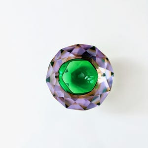 Op de afbeelding: Een facetgeslepen decoratief glazen object met een centraal groen, edelsteenachtig element. Het buitenste deel is lavendelkleurig met een geometrisch ontwerp. Het object staat tegen een witte achtergrond.