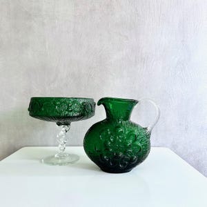 Può includere: Un set di calice e brocca in vetro verde. Il calice ha un bordo decorativo e un gambo trasparente e attorcigliato. La brocca ha un corpo rotondo, un manico e una superficie strutturata. Entrambi i pezzi sono verde smeraldo.