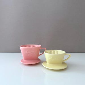 Könnte beinhalten: Zwei Keramik-Kaffeefilter, einer rosa und einer gelb, jeweils mit passender Untertasse. Der rosa Filter hat den Text "Melitta 101" auf der Vorderseite. Beide haben geriffelte Innenräume und kleine Griffe.