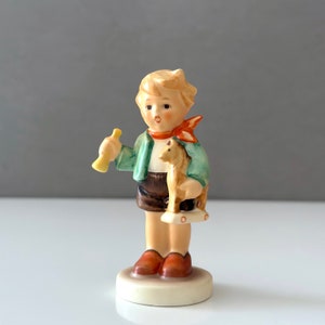 Könnte beinhalten: Eine Porzellanfigur eines Jungen, der ein Spielzeug-Hündchen und ein gelbes Horn hält. Der Junge trägt eine grüne Jacke, braune Shorts und einen roten Schal. Die Figur steht auf einem weißen Sockel.
