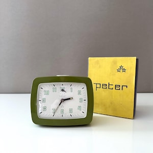 Puede incluir: Un reloj despertador vintage verde oliva con esfera blanca y manecillas negras. El reloj tiene forma rectangular con esquinas redondeadas y números blancos. El reloj está junto a una caja amarilla con el nombre "peter" impreso.
