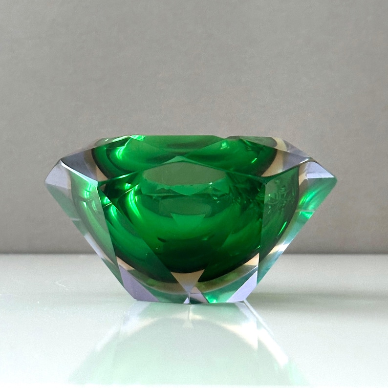 Op de afbeelding: Een facetgeslepen, smaragdgroene glazen kom met een geometrisch ontwerp. De kom heeft een diep, afgerond interieur en schuine zijkanten, die het licht reflecteren. Het is een decoratief item.