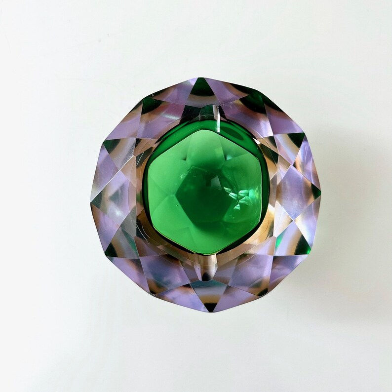 Op de afbeelding: Een facet asbak met een centrale groene glazen kom. De buitenrand is lichtpaars met een ruitpatroon. De asbak heeft een goudkleurige metalen rand. Het ontwerp is geometrisch en decoratief.
