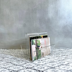 May include: A clear acrylic magazine holder with a curved top and a slanted front. The holder contains several magazines with colorful covers. The holder is sitting on a patterned rug.