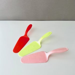 Puede incluir: Tres palas de pastel coloridas en rojo, verde claro y rosa. Cada pala tiene una hoja ancha y plana y un mango curvo. Las palas son de plástico y están dispuestas sobre una superficie blanca.