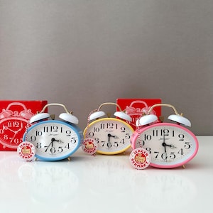 May include: A collection of colorful Jerger alarm clocks. The clocks have an oval shape with a white face, black numbers, and gold-colored bells. The clocks are in red, blue, yellow, and pink. Each clock has a small tag that says "Jerger King Time Germany."