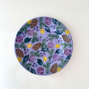 Peut inclure: Une assiette ronde en céramique avec un fond lavande, décorée d'un motif sur le thème de Noël. Le motif comprend des pommes de pin, des ornements, des feuilles de houx et des étoiles jaunes. L'assiette mesure environ 25 cm de diamètre.