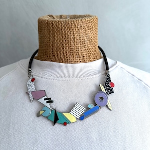 Puede incluir: Un collar geométrico colorido con un cordón negro. El colgante presenta formas en verde azulado, amarillo, morado y negro con detalles en rojo y plata. El collar se muestra sobre una camiseta blanca.