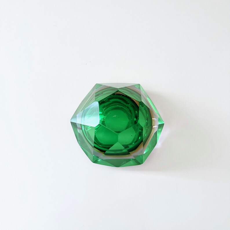 Op de afbeelding: Een gefacetteerde, smaragdgroene glazen asbak met een geometrisch ontwerp. De asbak heeft een zeshoekige vorm en staat tegen een witte achtergrond. De binnenkant van de asbak is een donkerdere tint groen.