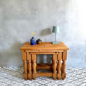 May include: A set of three wooden nesting tables with turned legs. The tables are made of light-coloured wood and have a square top. A small blue vase, a small yellow vase, and a green lamp sit on the top table. The tables are on a patterned rug.