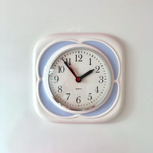 Könnte beinhalten: Eine quadratische Wanduhr mit abgerundeten Ecken, einem weißen Rahmen und einem hellblauen Innenrand. Das Zifferblatt ist weiß mit schwarzen arabischen Ziffern, schwarzen Stunden- und Minutenzeigern sowie einem dünnen roten Sekundenzeiger. QUARTZ steht darauf.