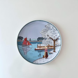May include: A ceramic plate featuring a winter scene. The scene shows a sailboat on a lake with a house and a woman standing on the shore. The trees are covered in snow and the sky is a pale blue.