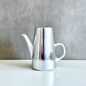 May include: A modern, silver-colored metal teapot with a white, curved spout and handle. The teapot has a conical shape and sits on a white surface. The background is a textured gray wall. The teapot is designed for serving beverages.