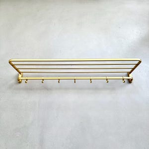 May include: A gold-colored metal wall-mounted shelf with a rack and seven hooks. The shelf has a rectangular design with four horizontal bars and a curved edge. The hooks are evenly spaced along the bottom.