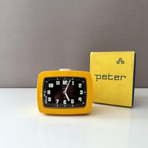 Puede incluir: Un despertador amarillo vintage con esfera negra y números blancos. El reloj tiene forma rectangular con esquinas redondeadas y un botón blanco en la parte superior. Una caja amarilla con el nombre "peter" está junto al reloj.