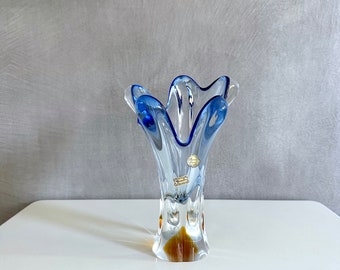 May include: A clear glass vase with a blue rim and a brown base. The vase has a wavy, organic shape and is decorated with a small label.