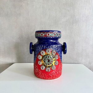以下が含まれることがあります： 青いセラミック花瓶。表面はテクスチャ加工で、両側にハンドルが2つ付いています。赤色の円形デザインで、金と緑のアクセントがあります。上部の縁には赤、青、金色の装飾模様があります。