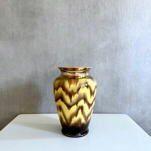 Könnte beinhalten: Eine gelbe und braune Keramikvase mit einem wellenförmigen Muster. Die Vase hat eine glänzende Oberfläche.