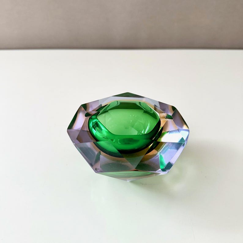 Op de afbeelding: Een facet geslepen, decoratieve glazen asbak met een centrale groene kom. De asbak heeft een geometrisch ontwerp met paarse en heldere glazen facetten. De binnenkom is een levendige groene kleur.