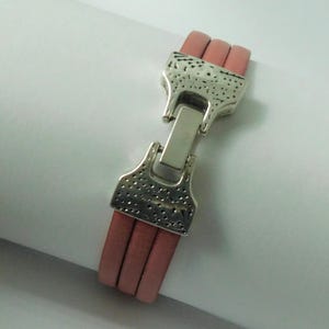 Puede incluir: Una pulsera que presenta tres cordones de cuero rosa polvoriento paralelos asegurados por un cierre de metal plateado. El cierre tiene una superficie texturizada y martillada con un mecanismo de bloqueo rectangular central que une los extremos.