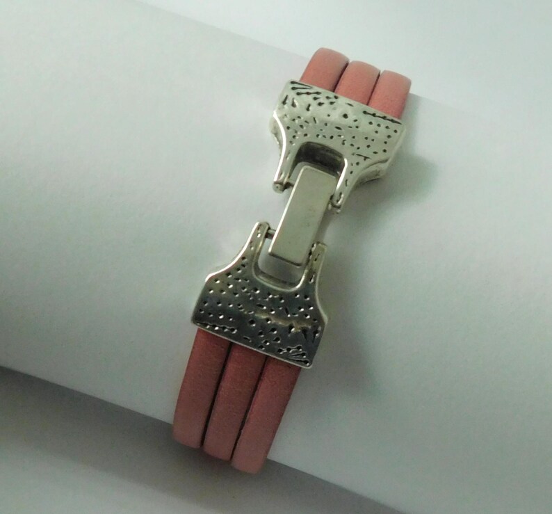 Puede incluir: Una pulsera que presenta tres cordones de cuero rosa polvoriento paralelos asegurados por un cierre de metal plateado. El cierre tiene una superficie texturizada y martillada con un mecanismo de bloqueo rectangular central que une los extremos.