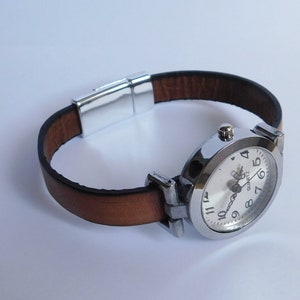 May include: A silver-toned wristwatch with a white face and black numbers. The watch has a brown leather band with a magnetic clasp.