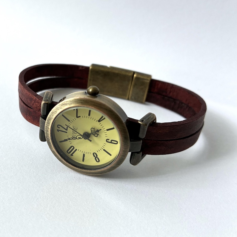 May include: A wristwatch featuring a round, antique brass-toned case with a pale yellow dial, black numerals, and ornate hands. The band consists of two parallel dark brown leather straps connected by a matching brass-toned magnetic clasp.