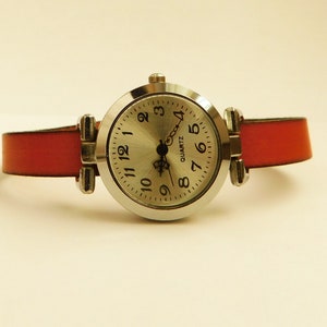 May include: A silver-toned quartz watch with a round face and a red leather band. The watch face has black numerals and hands. The band is attached to the watch case with silver-toned metal hardware.