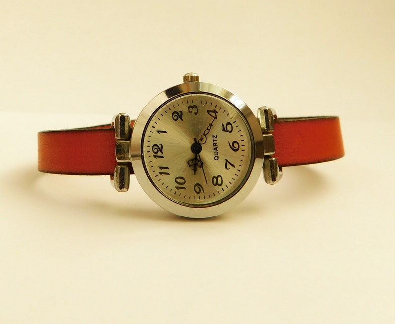 May include: A silver-toned quartz watch with a round face and a red leather band. The watch face has black numerals and hands. The band is attached to the watch case with silver-toned metal hardware.