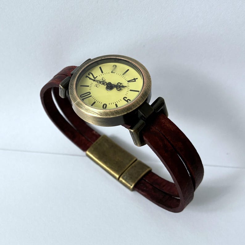 May include: A wristwatch featuring a round, antique brass-toned metal case with a pale yellow dial and black Arabic numerals. The watch has ornate black hands and a double-strand dark maroon leather strap secured by a rectangular brass-toned magnetic clasp.