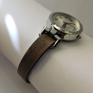 May include: A silver wristwatch with a brown leather band. The watch face has black Roman numeral hour markers and a white background.