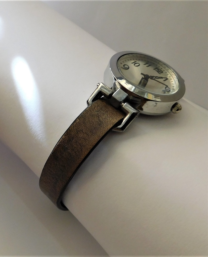 May include: A silver wristwatch with a brown leather band. The watch face has black Roman numeral hour markers and a white background.