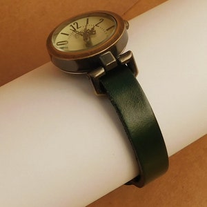 Op de afbeelding: Een rond, antiek-stijl horloge met een cr&egrave;mekleurige wijzerplaat en zwarte Romeinse cijfers. Het horloge heeft een donkergroene leren band.