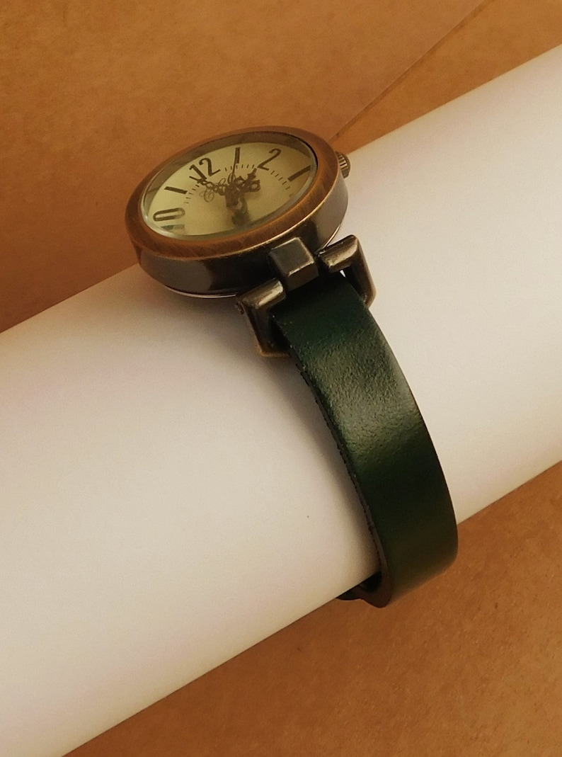 Op de afbeelding: Een rond, antiek-stijl horloge met een cr&egrave;mekleurige wijzerplaat en zwarte Romeinse cijfers. Het horloge heeft een donkergroene leren band.