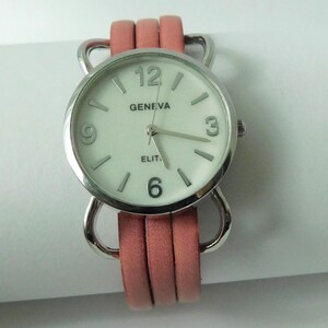 Puede incluir: Un reloj de pulsera con caja redonda plateada y esfera blanca con el texto Geneva y Elite. El reloj tiene marcadores de hora plateados, n&uacute;meros ar&aacute;bigos en el 3, 6, 9 y 12, y una correa de tres hilos de color coral unida a la caja.