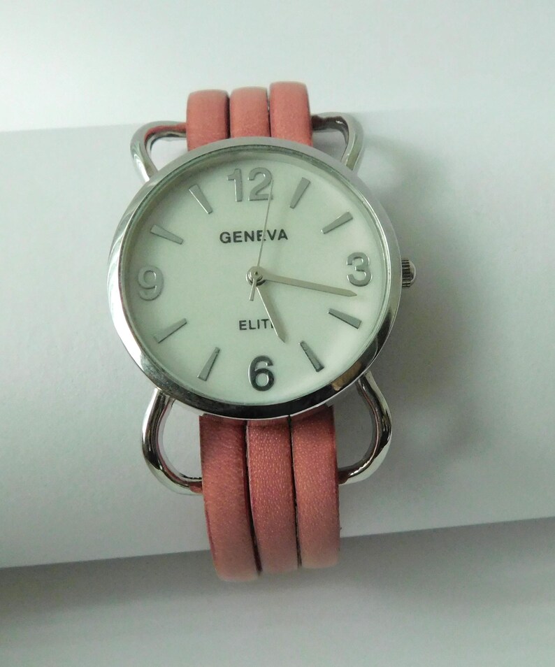 Puede incluir: Un reloj de pulsera con caja redonda plateada y esfera blanca con el texto Geneva y Elite. El reloj tiene marcadores de hora plateados, n&uacute;meros ar&aacute;bigos en el 3, 6, 9 y 12, y una correa de tres hilos de color coral unida a la caja.