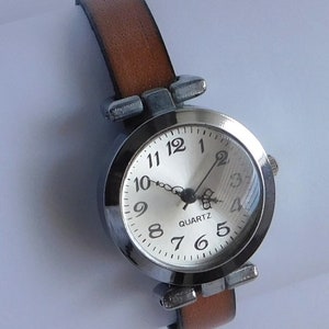 May include: A silver-toned wristwatch with a brown leather band. The watch face is white with black numbers and hands. The watch face also has the word "QUARTZ" printed on it.