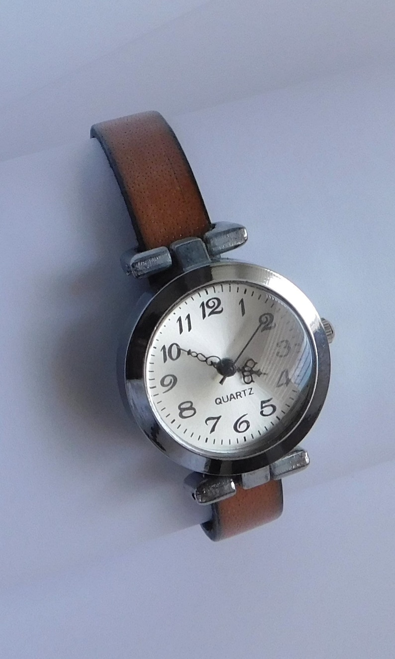 May include: A silver-toned wristwatch with a brown leather band. The watch face is white with black numbers and hands. The watch face also has the word "QUARTZ" printed on it.