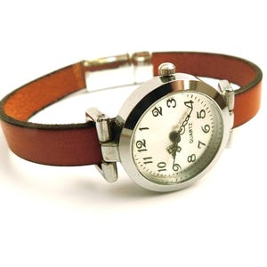 May include: A silver quartz watch with a white face and black numerals. The watch has a brown leather band. The watch face is round and the band is thin. The watch is on a white background.