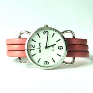 Puede incluir: Un reloj de pulsera con caja redonda plateada y esfera blanca con el texto Geneva Elite. El reloj tiene manecillas negras para horas, minutos y segundos, n&uacute;meros negros en el 3, 6, 9 y 12, y una correa de cuero rosa de varias hebras con bucles plateados.