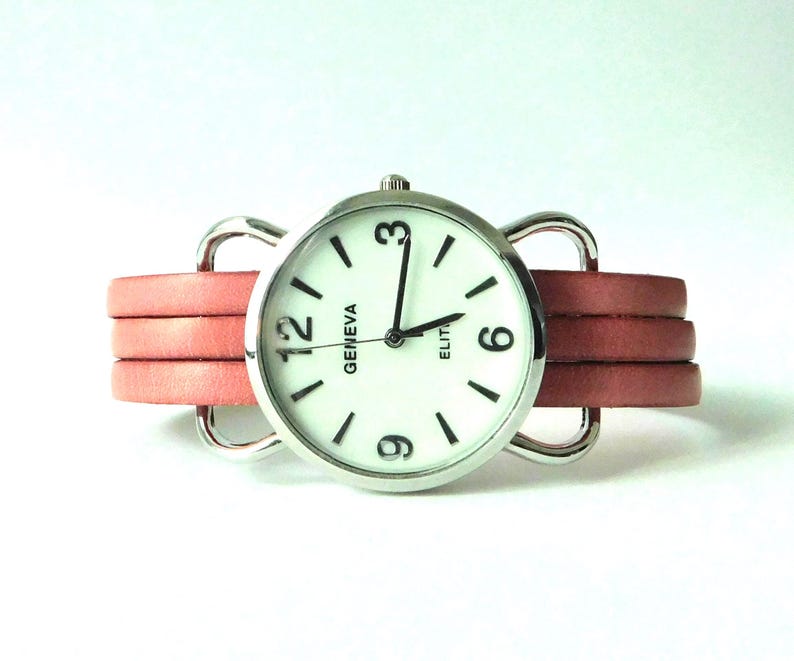 Puede incluir: Un reloj de pulsera con caja redonda plateada y esfera blanca con el texto Geneva Elite. El reloj tiene manecillas negras para horas, minutos y segundos, n&uacute;meros negros en el 3, 6, 9 y 12, y una correa de cuero rosa de varias hebras con bucles plateados.