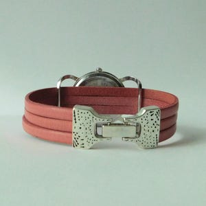 Puede incluir: Un reloj de pulsera con una correa de cuero rosa de tres hilos y un cierre de metal plateado con un acabado texturizado y moteado. La caja del reloj es circular con una parte posterior plateada, situada detr&aacute;s de la correa.
