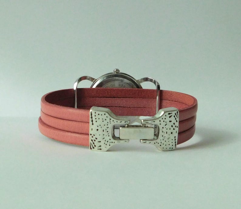 Puede incluir: Un reloj de pulsera con una correa de cuero rosa de tres hilos y un cierre de metal plateado con un acabado texturizado y moteado. La caja del reloj es circular con una parte posterior plateada, situada detr&aacute;s de la correa.