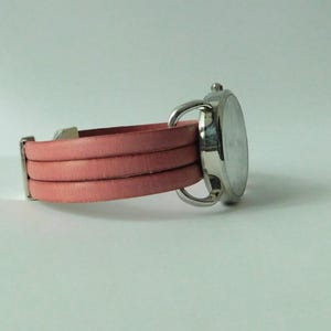 Puede incluir: Un reloj de pulsera con caja redonda de metal plateado y correa de varias vueltas hecha de cuero rosa liso. La correa consta de tres bandas distintas que se conectan a la caja del reloj con herrajes de metal plateado y una peque&ntilde;a corona.
